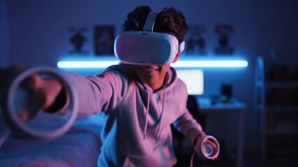 A young man wearing a virtual reality headset and holding controllers experiences augmented reality in a dark room with blue lighting. - Powered by Adobe