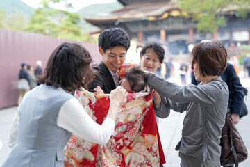 A group of people are gathered around a baby wearing a red and white kimono. The baby is being held by a woman in a gray jacket. Scene is joyful and celebratory