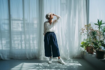 A senior woman stands gracefully in sunlight, surrounded by indoor plants, creating a serene and contemplative atmosphere.