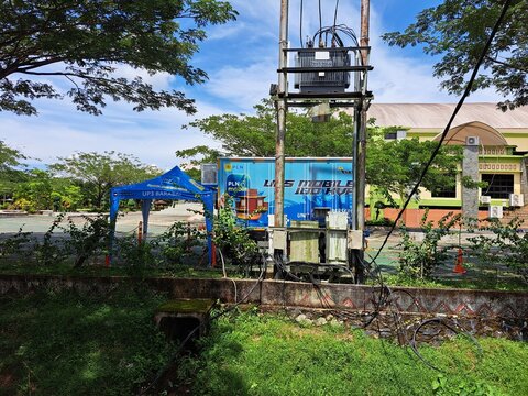 Tapin, Indonesia December 1st 2025 : ​PLN mobile UPS unit, likely used for temporary power backup or emergency supply, is set up outdoors in a grassy area with a canopy and building in the background