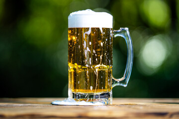 Golden craft beer poured outdoors. Beer splash against green nature background. Pouring amber ale in nature. Brewed beer with bubbles and foam. Craft brewery lager beer on summer day.