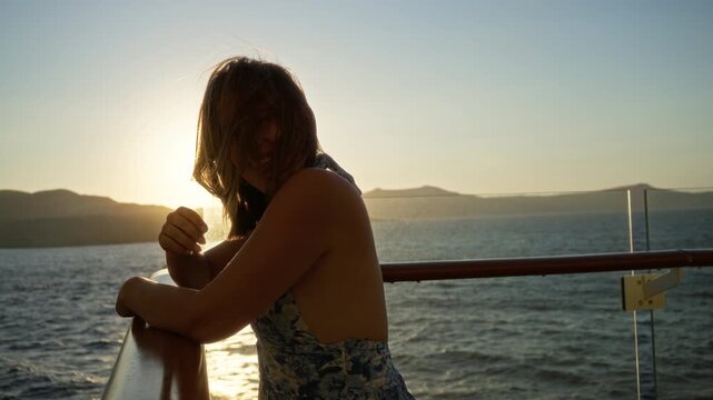 Woman leaning on cruise ship deck rail with bare back visible, hair blowing and smiling toward sunset over sea; carefree joy.