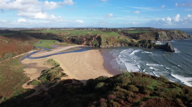 Drone gliding above a sandy beach, rugged cliffs with waves below and grassy headlands beyond at Three Cliffs Bay on the Gower Peninsula in Wales