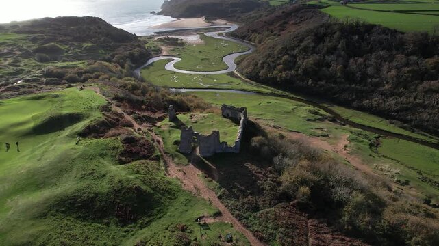 Drone circling the ruins of Pennard Castle while revealing Three Cliffs Bay valley and winding Pennard Pill river in Wales