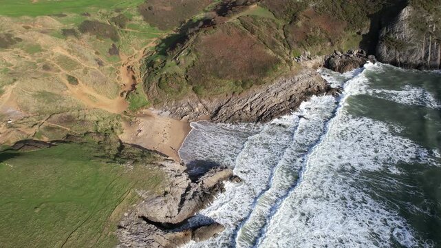 Circling view over Pobbles Bay with foaming waves rocks and coastal grasslands along the Gower Peninsula shoreline in Wales