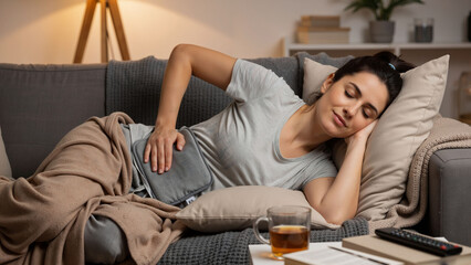 Obraz premium Woman relaxing on sofa with heating pad on her stomach. Female suffering from abdominal pain or menstrual cramps on comfortable couch. Home therapy.