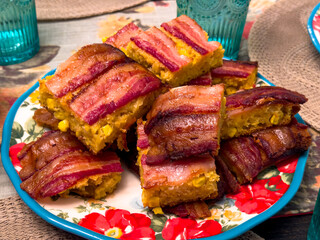 Close-up of bacon-topped cornbread casserole bars stacked on a floral plate. Warm colors and textures make it ideal for comfort food, holiday meals, potlucks, and recipe themes.