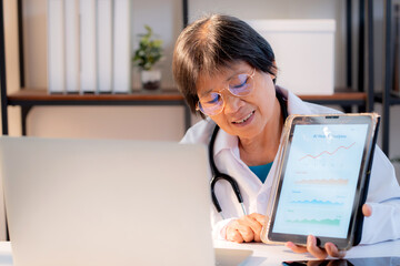 Senior asian doctor presenting AI health analysis on tablet during consultation with patient in clinic, healthcare technology, elderly female physician explaining health data chart, medical concept.