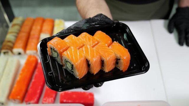 A close-up chef in gloves holds a disposable plate of sliced sushi in the restaurant's kitchen. The process of making and serving sushi in a restaurant.