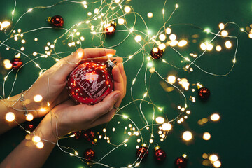 Female hands holding red Christmas ornament on green background with Christmas light garland,...