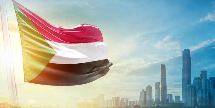Sudan flag wavering in beautiful clouds with building skyline. The flag with drone shot in beautiful sky. Sudan national flag for independence day.
