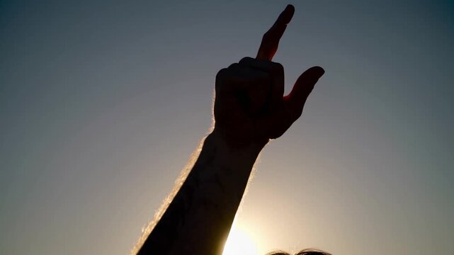 A hand is raised toward the sky, fingers pointed upward in a gesture of triumph against a stunning sunset backdrop. The light creates a silhouette, emphasizing the strength and det