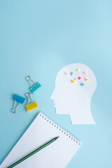 A paper cutout of a human head with colorful sticky notes on a blue background. Nearby are binder clips and a notepad with a pencil.