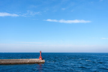 A small, vibrant red lighthouse structure stands on a concrete pier, contrasting with the deep blue ocean waves and a clear sky above.
