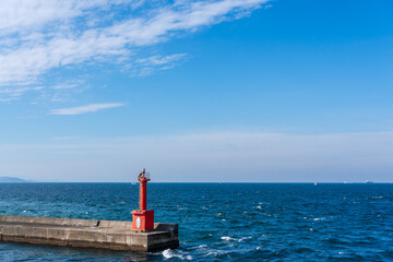 A red navigation beacon sits at the end of a solid breakwater pier, set against the vast blue expanse of the sky and the open sea.