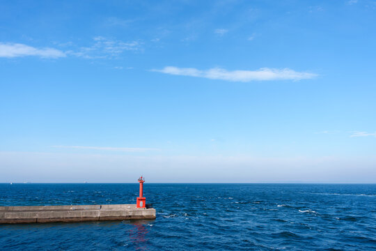 A small, vibrant red lighthouse structure stands on a concrete pier, contrasting with the deep blue ocean waves and a clear sky above.
