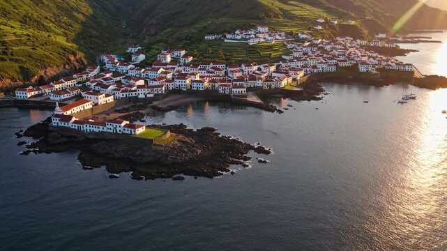 Charming white houses with red roofs sit along the rocky coastline, creating a picturesque village by the sea. Verdant hills rise in the background, framing the idyllic coastal sce
