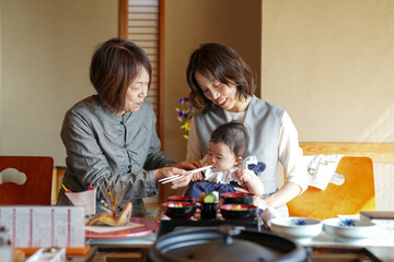 A woman is feeding a baby with chopsticks. The baby is wearing a blue dress. The woman is holding the baby's hand while feeding it