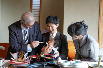 A man feeds a baby while a woman watches. The baby is wearing a white dress. The family is gathered around a dining table with food and drinks