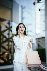 Fototapeta premium An Asian woman happily talking on the phone while holding shopping bags outside a modern building, capturing the joyful lifestyle of a young Asian woman on a shopping day.