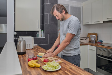 A man slices bread and assembles ingredients on a wooden counter while preparing a homemade sandwich.