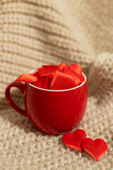 A red mug is filled with red heart-shaped confetti sitting on beige knitted fabric. Two red hearts lay next to the mug. Suitable for Valentine's Day digital content.