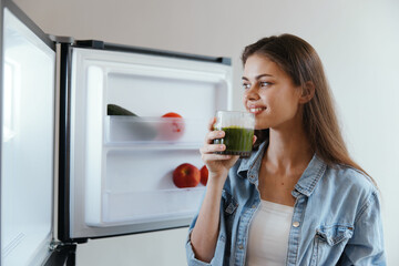 Healthy woman enjoying green smoothie in front of open fridge filled with fresh fruits and...