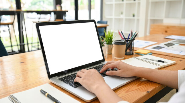A person's hands typing on a laptop computer placed on a wooden desk with notebooks pens coffee cup and potted plant work office table technology business