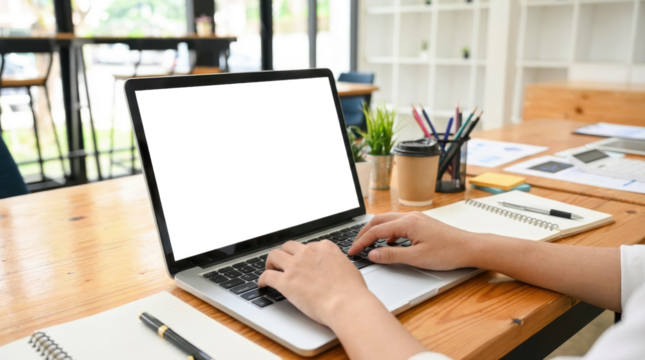 A person's hands typing on a laptop computer placed on a wooden desk with notebooks pens coffee cup and potted plant work office table technology business