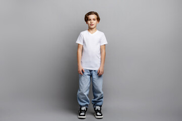 Full-length portrait of young boy standing in studio, wearing white t-shirt, trendy sneakers and jeans, looking at camera with relaxed expression on clean grey background