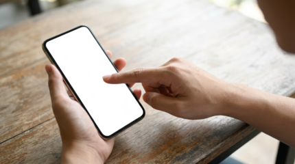 A person's hands holding and interacting with a modern smartphone on a wooden table with the screen transparent and ready for content mobile touching technology