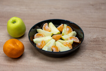 Apple slices and tangerine segments arranged in a black bowl on a wooden table, with whole apple and tangerine placed beside it. Fresh fruit snack prepared indoors.