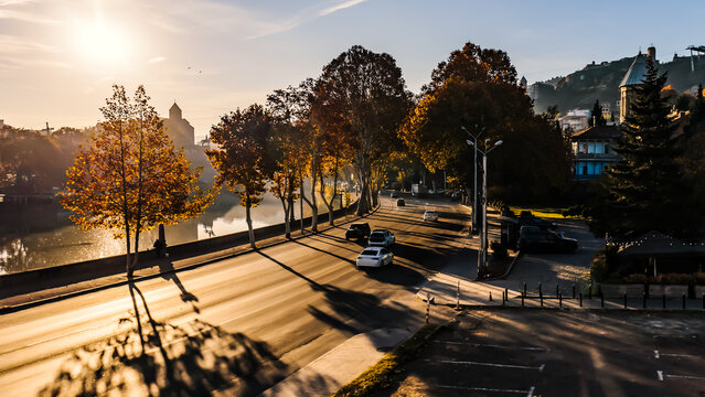 Tranquil autumn morning in Tbilisi, Georgia. Golden sunrise lights up a tree-lined avenue by the river as a few cars pass through the peaceful city scene