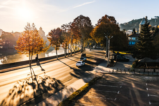 Morning sunlight casts long shadows over a quiet riverside street in Tbilisi, Georgia. Autumn trees glow in golden hues, reflecting on the calm surface of the water
