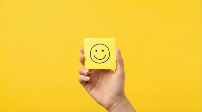 Hand holding yellow sticky note with smiley face illustration against vibrant yellow background, conveying positivity and cheerful emotions in a creative setting