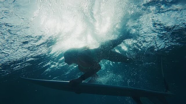 Woman surfer in bikini dives under the ocean wave with surfing board and passes it. Underwater view of the surfer doing duck dive trick