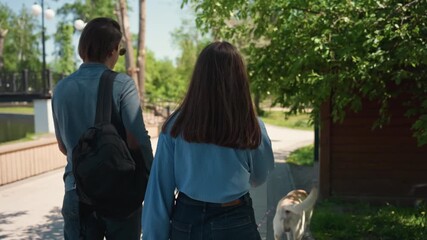 white couple walking past wooden pavilion through sunlit park, backpack and leash in hand, dog sniffing grass by pathway, leafy trees and warm summer day framing leisurely neighborhood walk