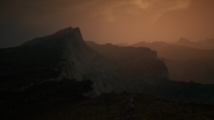 Mountains rise dramatically against a darkening sky, bathed in warm golden light from the setting sun. Mist envelops the rugged terrain, creating an air of mystery and tranquility.