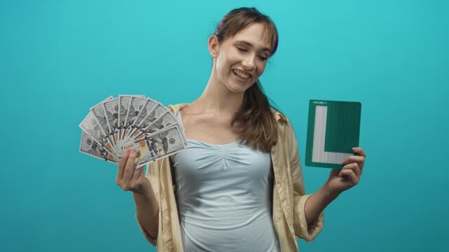 Young woman smiling and holding a fan of us dollar bills and a learner l plate in a turquoise studio; confidence.