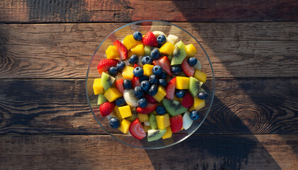 A clear glass bowl filled with a vibrant fruit salad featuring blueberries, mango, kiwi, and strawberries, resting on a rustic wooden table under natural light.