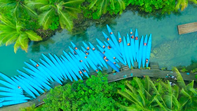 Aerial view of numerous blue kayaks docked at a wooden pier on a clear river, surrounded by lush tropical vegetation with people in life vests, Maasin River, Siargao, Philippines.