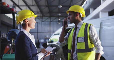 Discussing two wearing hard hats and hi-vis vest with clipboard, speaking into radio in workshop