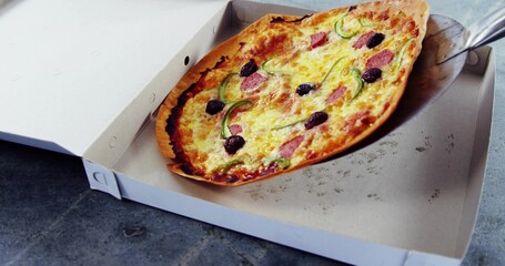 Fototapeta premium Lifting thin-crust pizza on metal spatula in white box on gray tabletop, showing pepperoni, olives