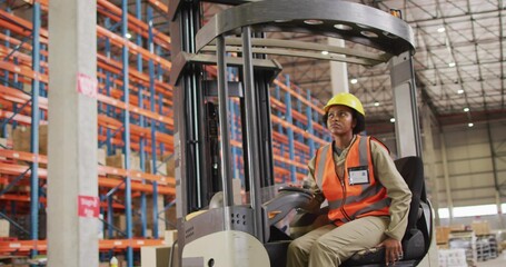 Operating forklift worker with yellow hardhat orange vest steering past pallet racking, copy space
