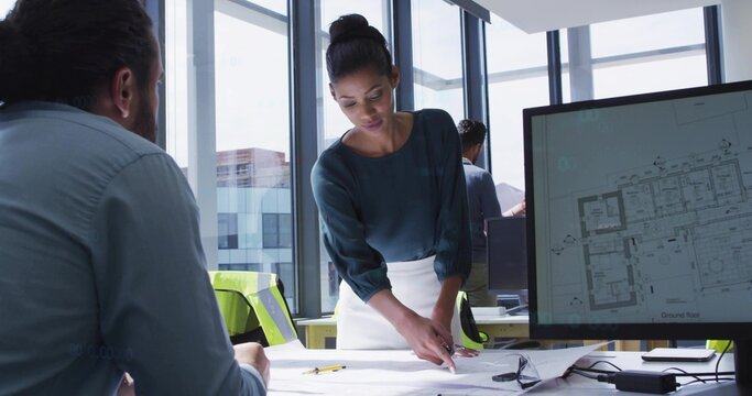 Pointing lead in teal blouse, white skirt leaning over table in office with monitor, vest, plans