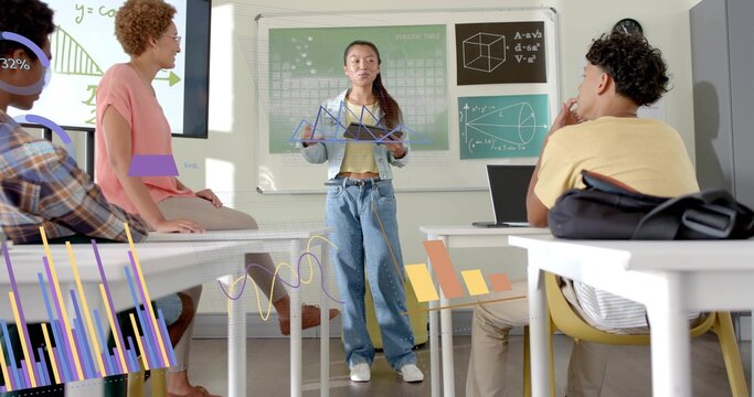 Presenting student in denim jacket holding triangle model at classroom front, with laptop graphs