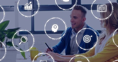 Reviewing colleagues in yellow cardigan blue sweater gesturing at laptop in office, analytics icons