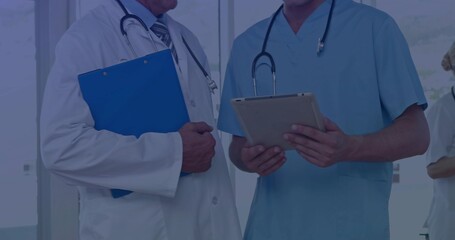 Standing in clinic coat-tie doctor holding blue clipboard scrubs clinician using tablet stethoscope
