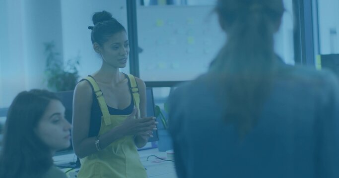 Standing woman in yellow overalls addressing team in open office, monitors and sticky notes