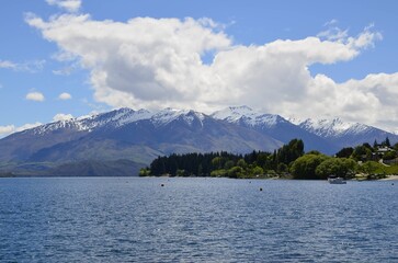 Snowy mountains reflecting over a blue lake in New Zealand, framed by forests and a cloudy sky, showcasing a majestic natural landscape.